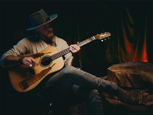 Jackson Dean sitting on a chair with a cowboy hat on playing the guitar